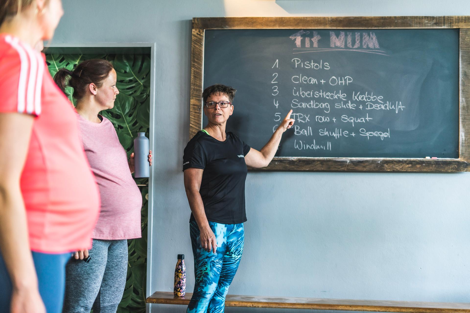 Fitnesstrainerin erklärt Kursteilnehmerinnen Übungsausführung an Tafel