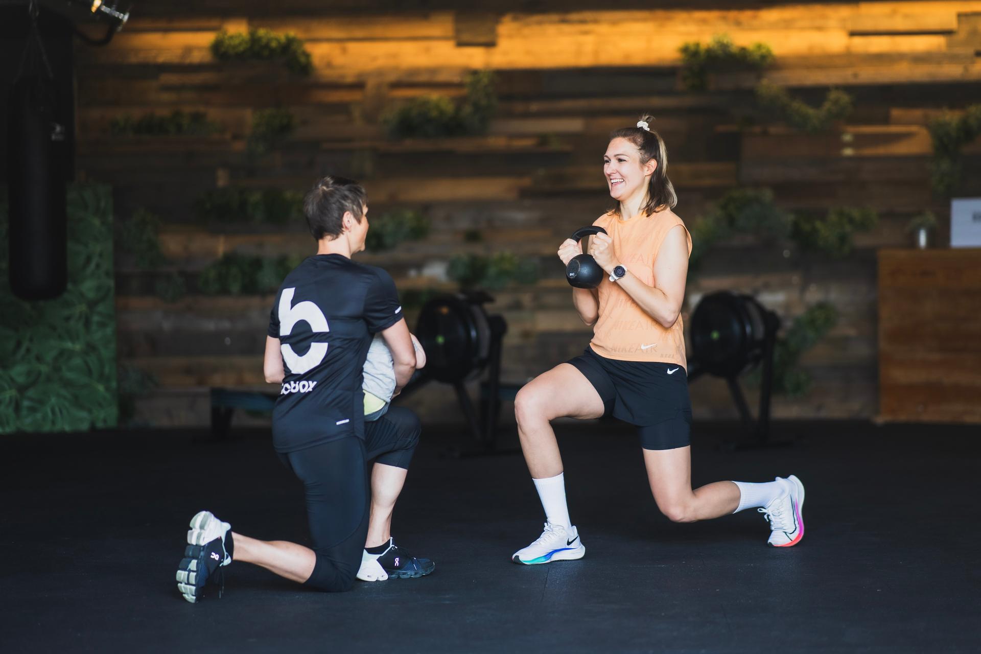 Trainerin mit Baby auf dem Arm zeigt Kurskundin Ausfallschritt mit Kettle Bell